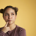 Brunette young woman touches her chin and looks up with a questioning expression against a yellow background