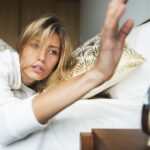 A blonde woman sleepily turns off her alarm clock