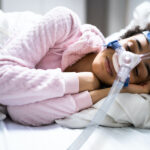 A Black woman wearing a CPAP machine as she sleeps on her side in her bed