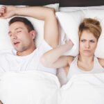 Aerial view of an irritated blonde woman in bed covering her ears with a pillow lying next to a snoring brunette man
