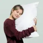 A blonde woman in a maroon sweater hugs her pillow while standing in front of a light green background